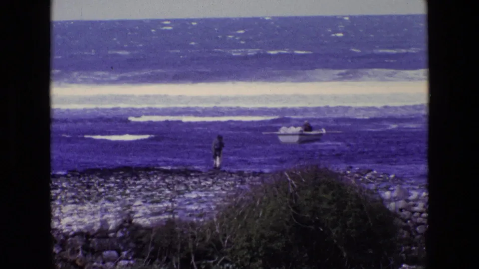 1969 8mm Ireland shoreline view with a distant person, small boat, rocky ground, coastal grass, and bright surf bands.