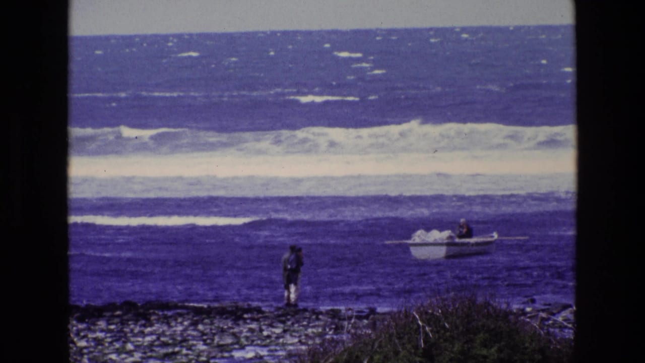 A distant person on an Irish rocky shoreline faces a small boat in rolling blue water, filmed on 8mm in 1969.