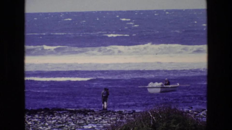 A distant person on an Irish rocky shoreline faces a small boat in rolling blue water, filmed on 8mm in 1969.