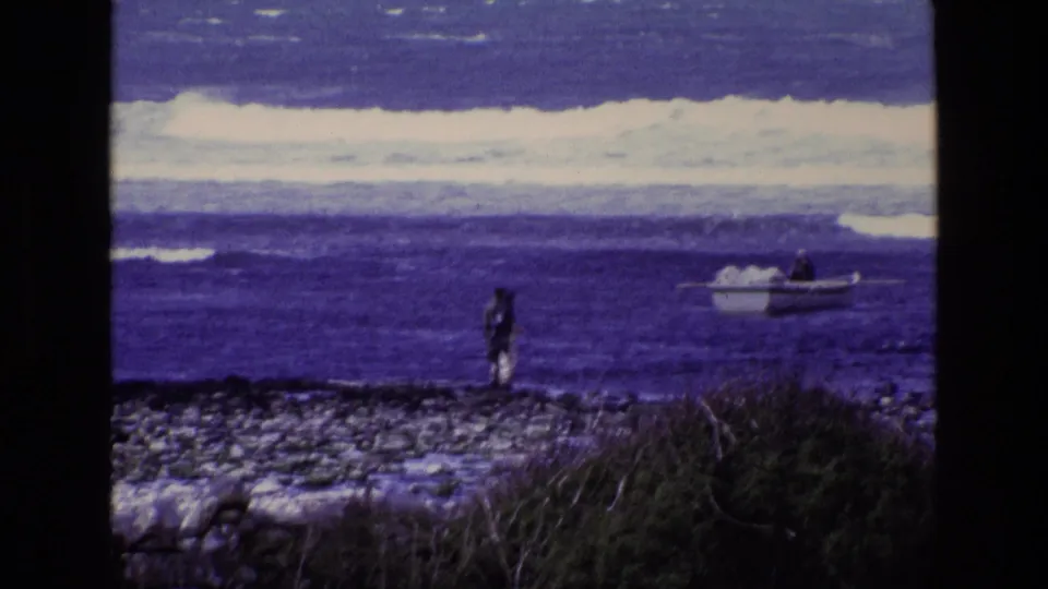 A 1969 Ireland coastal home-movie frame with a distant standing figure, low rocks, and a small boat near surf.