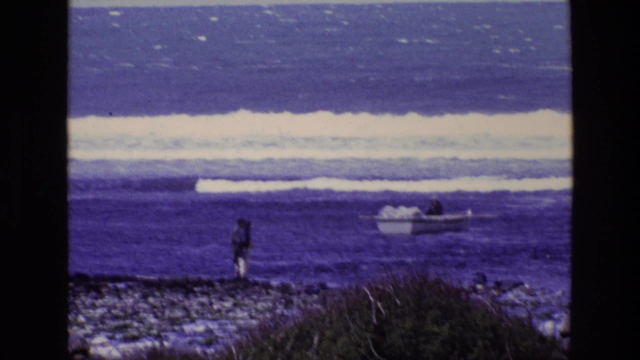 1969 Ireland coastal home-movie frame with a distant person facing the ocean and a small boat near breaking waves.