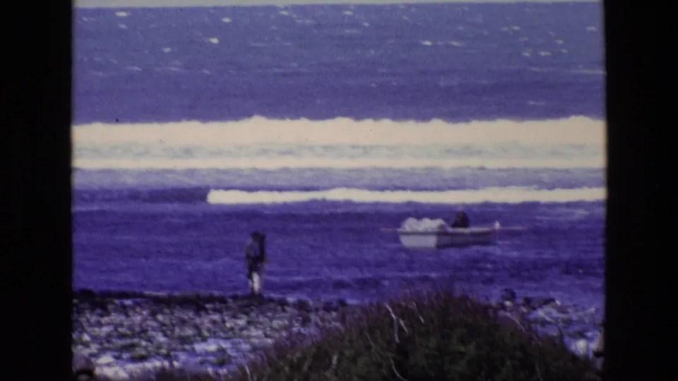 1969 Ireland coastal home-movie frame with a distant person facing the ocean and a small boat near breaking waves.