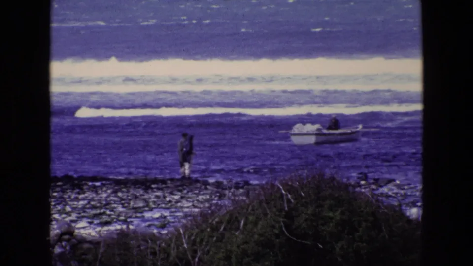 1969 Ireland 8mm still of a lone figure on a rocky beach looking toward a small boat in bright blue ocean water.