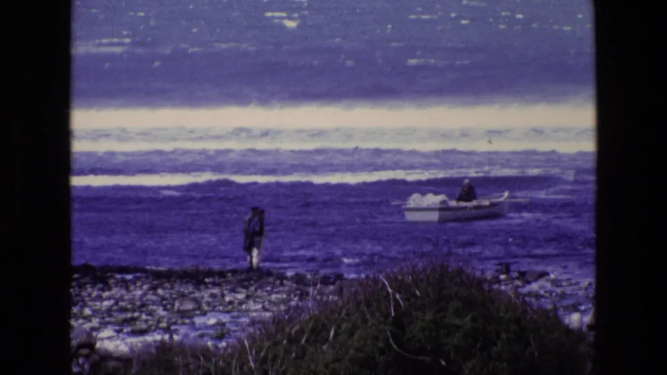 1969 Ireland coastal frame showing a shoreline figure, foreground brush, and a small open boat on blue water near surf.