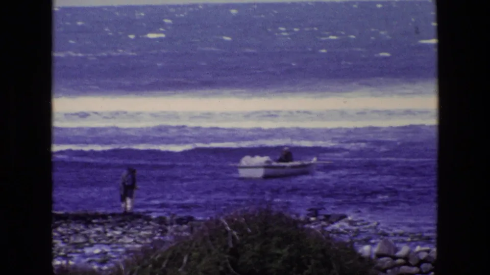 1969 Ireland 8mm frame showing a distant person on a rocky shoreline watching a small boat near breaking waves.