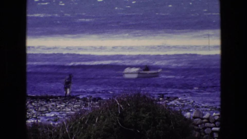 1969 Ireland coastal home-movie frame with a standing figure on rocks and a small boat floating just beyond the shore.