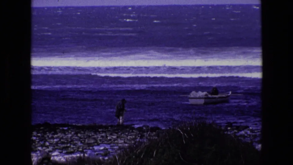 1969 Ireland 8mm frame showing a distant shore figure, a small open boat, dark rocks, brush, and layered surf.
