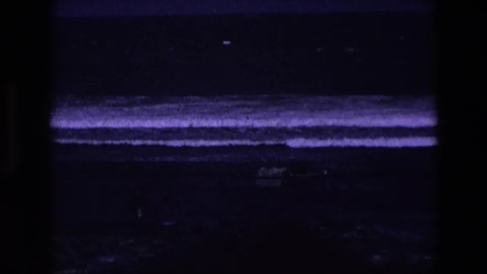 Underexposed 1969 Ireland coastal 8mm frame with visible surf bands, rocky foreground, and a faint small boat near the lower center.