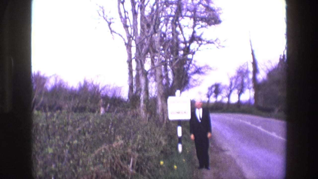 Man in a dark suit stands beside a Donegal road sign on a rural roadside.