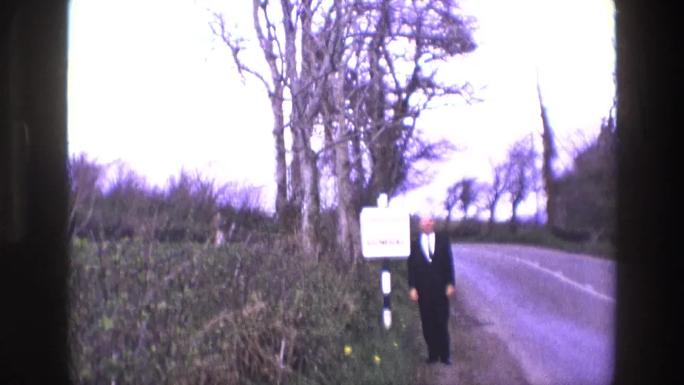 In rural Ireland, a man in a dark suit stands on a verge beside a bilingual Donegal sign near a paved road.