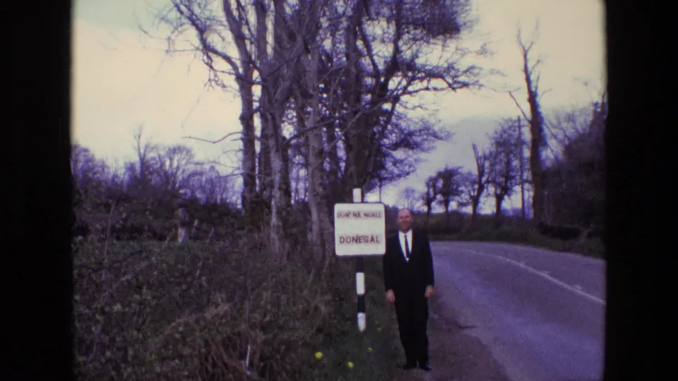 1969 8mm frame of a suited man beside the Dún na nGall and Donegal sign on a rural Irish roadside.
