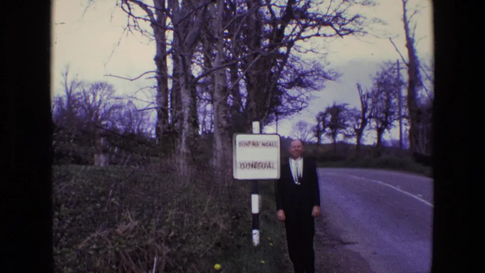 A suited man stands beside the Donegal road sign in a rural Irish landscape, with hedges and leafless trees behind.