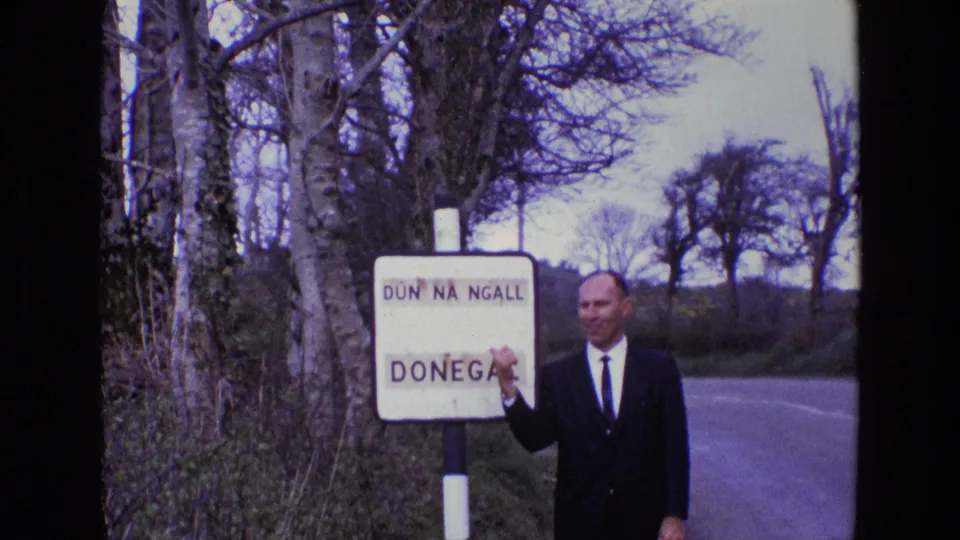 The suited man points at the Donegal sign, with bare trees and a rural road visible behind him.