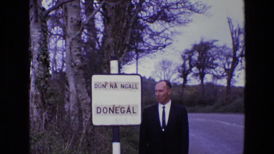 A suited man stands next to the Donegal sign and turns slightly away from the camera on a rural Irish roadside.