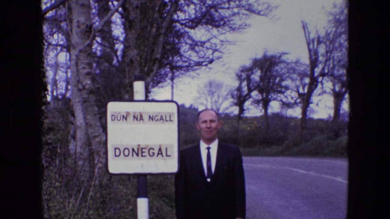 Man in a dark suit stands beside a Donegal road sign on a rural road.