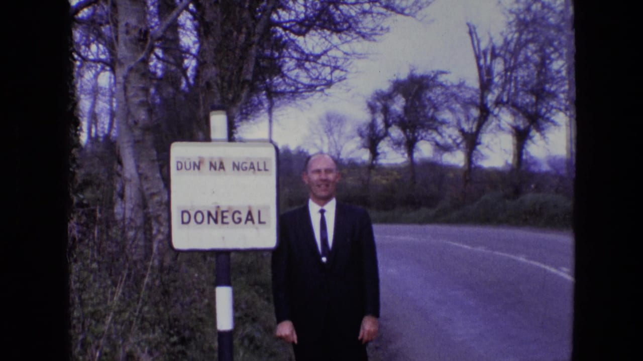 A smiling man in a dark suit poses beside the Donegal sign on a curving rural road in Ireland, supported by the clip metadata.
