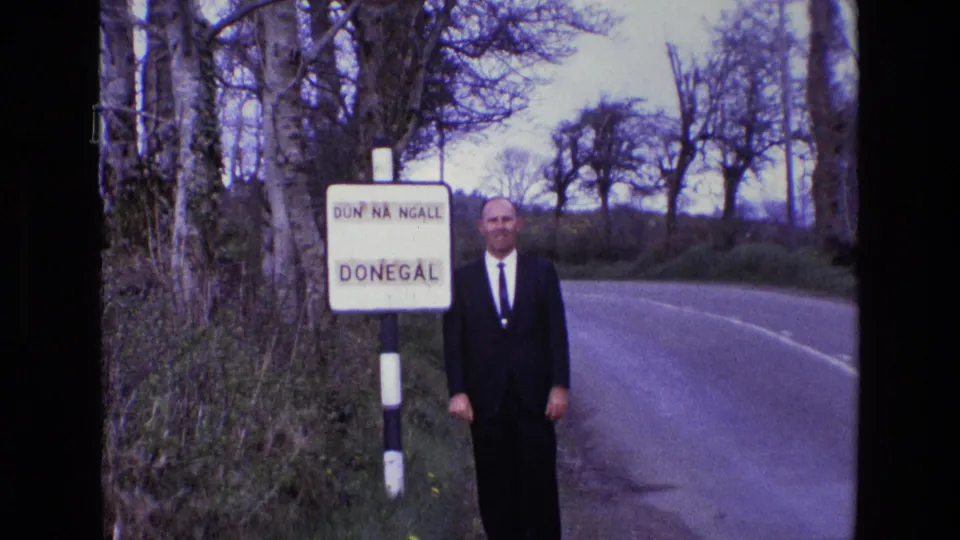 The wider 8mm frame shows the man standing full-length near the Donegal sign, with trees and a rural road behind him.