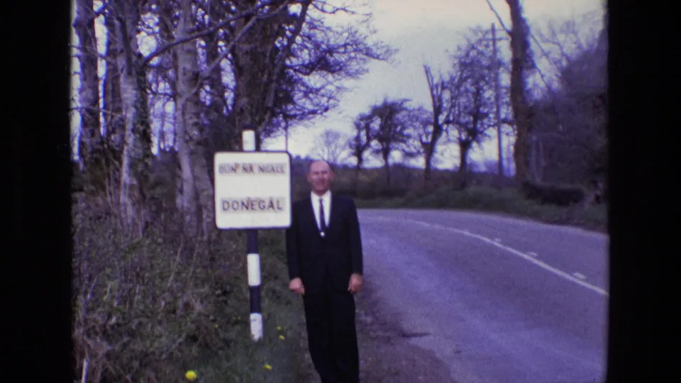 A wide 1969 Ireland travel frame shows the suited man beside the Donegal sign, with a curving road and bare trees in the background.
