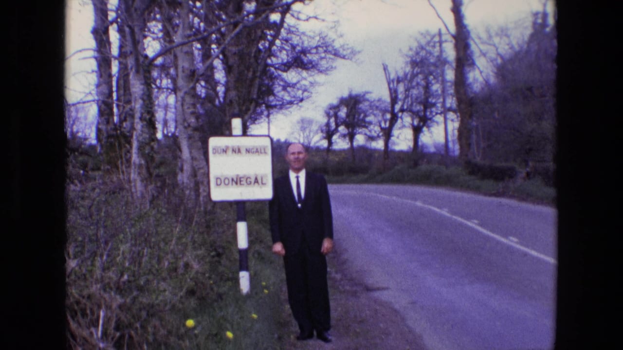 In 1969 Ireland, a man in a dark suit and tie stands near a bilingual Donegal sign beside a curving rural road lined with bare trees.