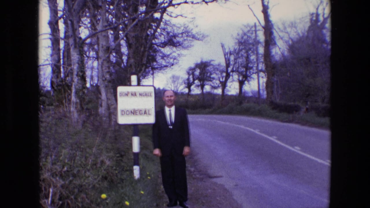 The suited man remains beside the bilingual Donegal roadside sign, facing the camera with the rural road and leafless trees behind him.
