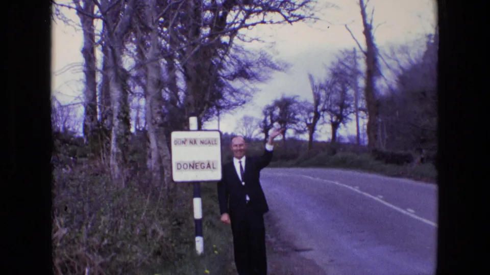 In the Ireland roadside scene, the suited man raises his hand in greeting while standing beside the Donegal sign on the grass verge.