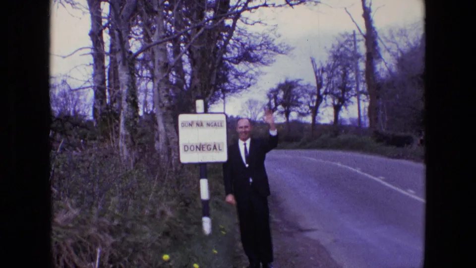 The suited man waves at the camera beside the bilingual Donegal sign, with a quiet rural road and winterlike trees in the background.
