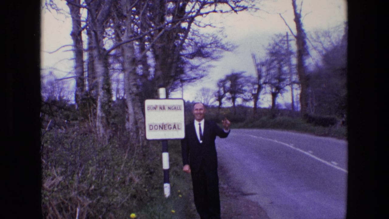 In 1969 Ireland, a suited man waves near a bilingual Donegal sign on a country road lined with trees and hedges.