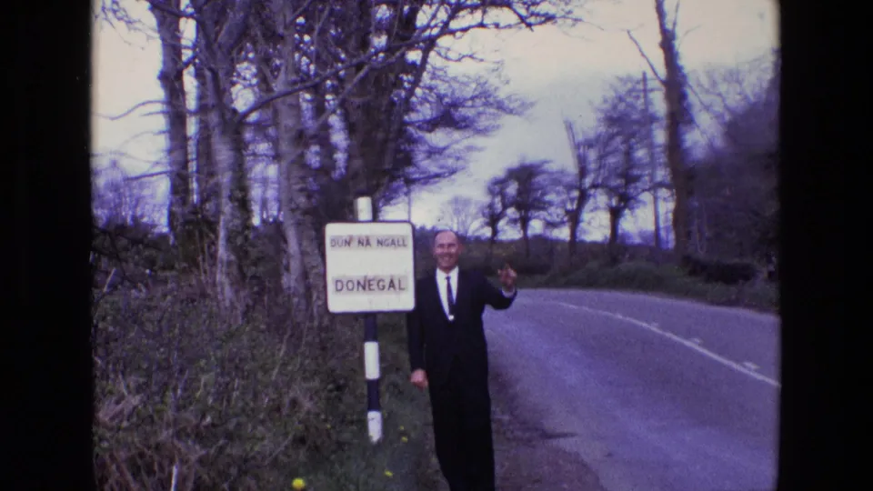 In 1969 Ireland, a suited man waves near a bilingual Donegal sign on a country road lined with trees and hedges.