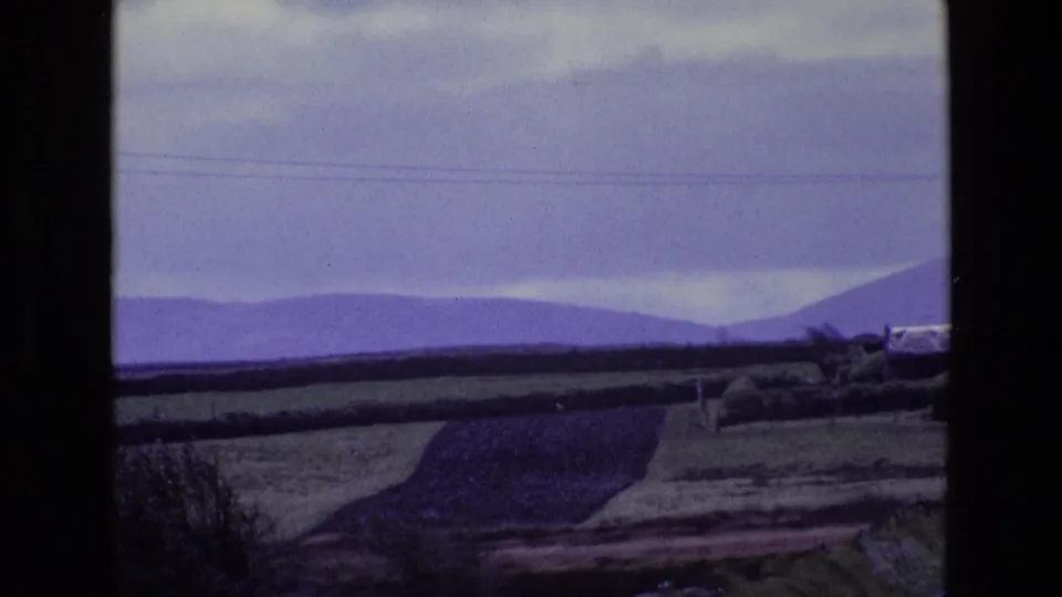 Rural Irish farmland stretches toward distant hills under a cloudy sky, with hedgerows and fields visible on 8mm film.