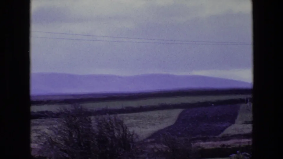 A 1969 Ireland countryside view shows patchwork fields, hedgerows, low hills, and overhead power lines beneath heavy cloud.
