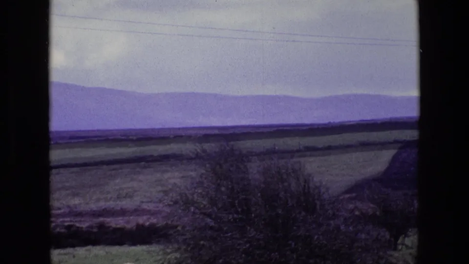 1969 8mm view of rural Ireland with green fields, hedgerows, distant purple mountains, clouds, and faint overhead power lines.