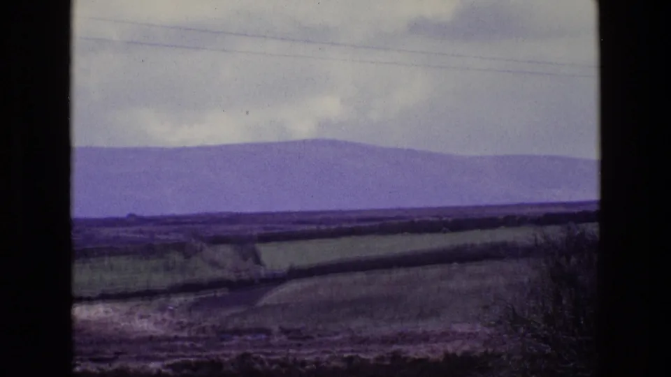 Wide 1969 8mm countryside view in Ireland, with fields divided by hedges, distant purple mountains, cloudy sky, and faint power lines.