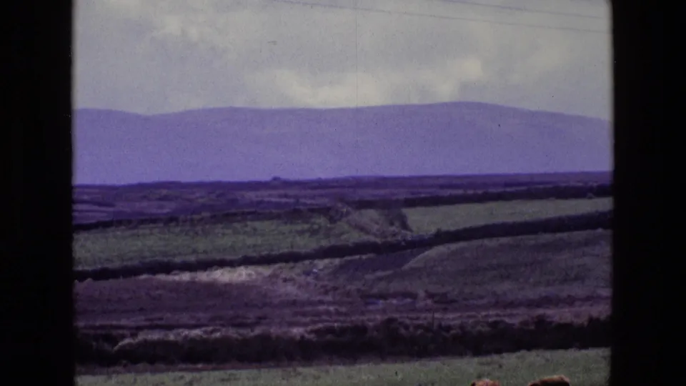 1969 rural Ireland frame with green fields, low hedgerows, distant purple mountains, cloud cover, faint power lines, and 8mm frame edges.