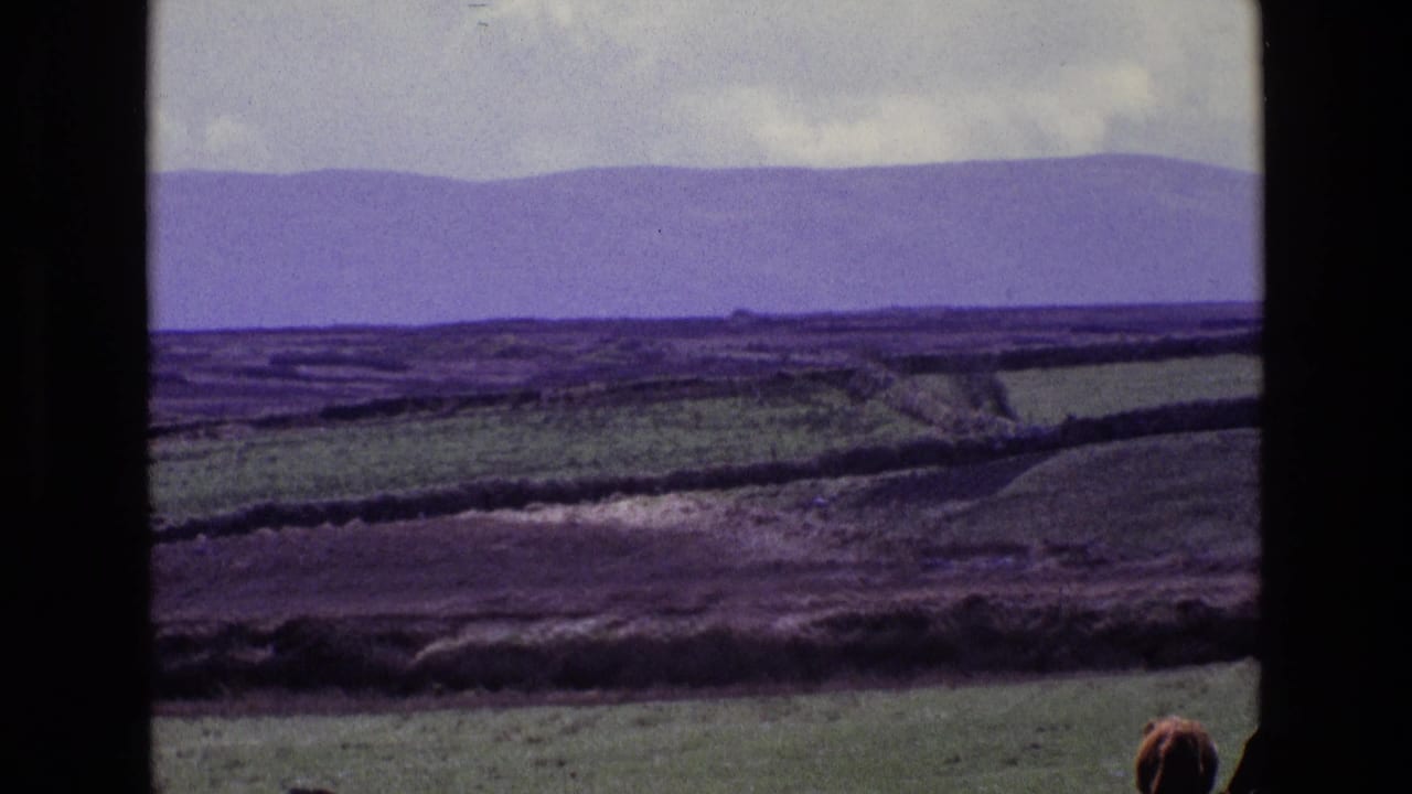 Rolling green and brown fields with distant hills under a cloudy sky, framed by dark vertical borders.