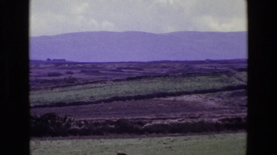 1969 Ireland home-movie frame of open countryside, layered field lines, distant hills, gray clouds, and dark framing at the sides.