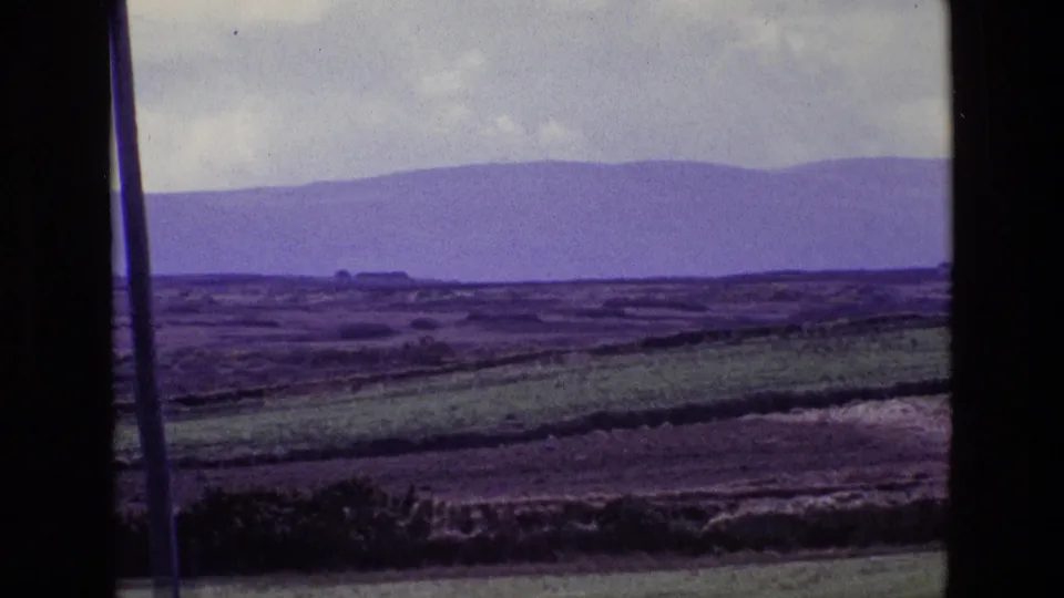 1969 Ireland 8mm frame with patchwork rural fields, hedgerow lines, distant purple hills, cloudy sky, and visible dark border framing.