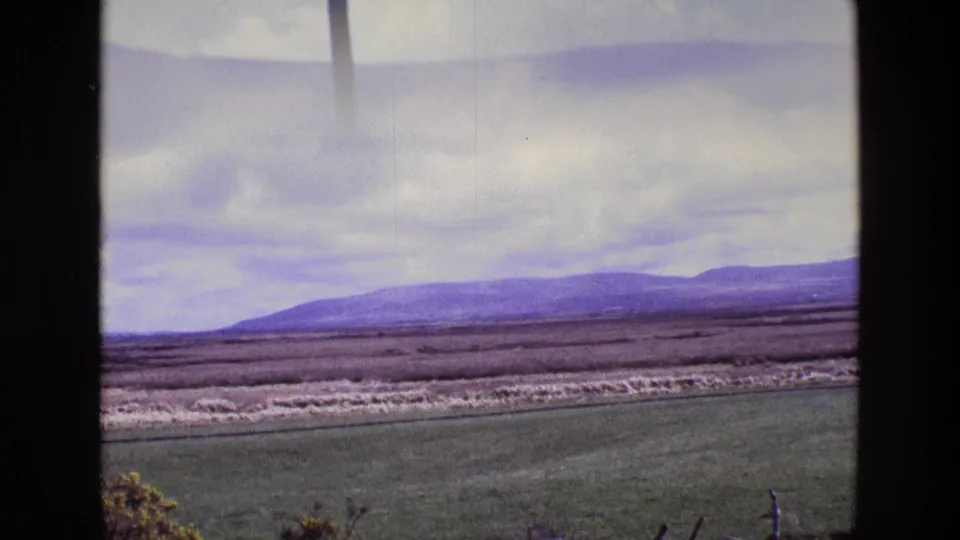 1969 Ireland 8mm frame showing a wider countryside view with foreground field, pale rough ground, distant hills, and overcast sky.