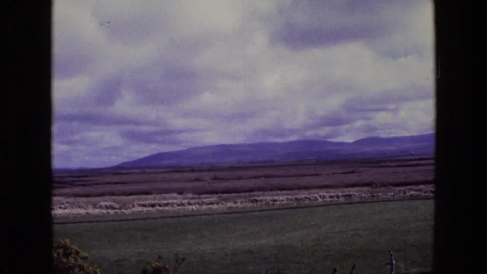 1969 Ireland 8mm frame showing rural fields, roadside vegetation, and a long mountain horizon beneath heavy cloud cover.