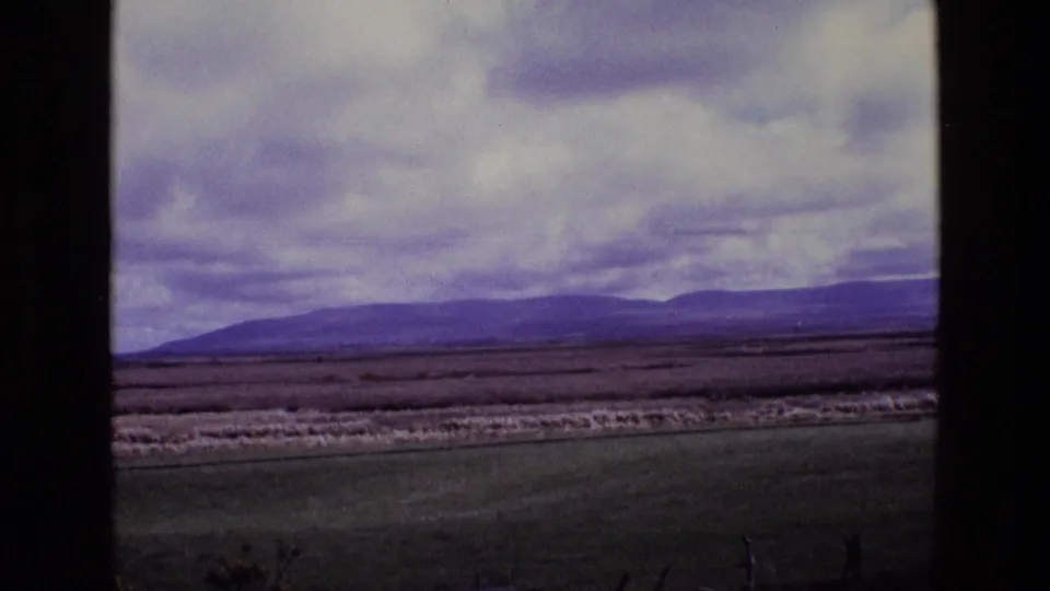 8mm home-movie frame from Ireland in 1969 showing a rural field view, low fence elements, and mountains beneath thick clouds.