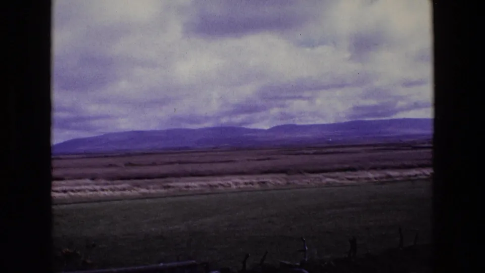 Late-1960s Ireland 8mm view across open fields toward a broad mountain range, with dark scan borders and muted color.