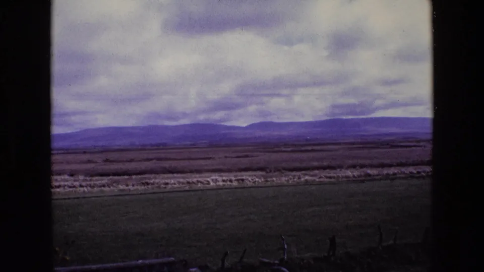 1969 Ireland home-movie frame showing green fields, low fencing, distant hills, and a cloud-filled sky.