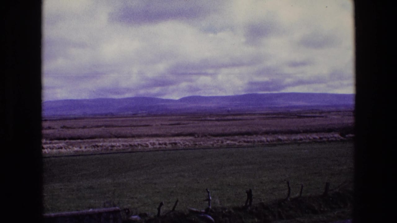 Wide 1969 Ireland countryside view with open fields, purple-toned hills, and dense clouds within dark 8mm frame edges.