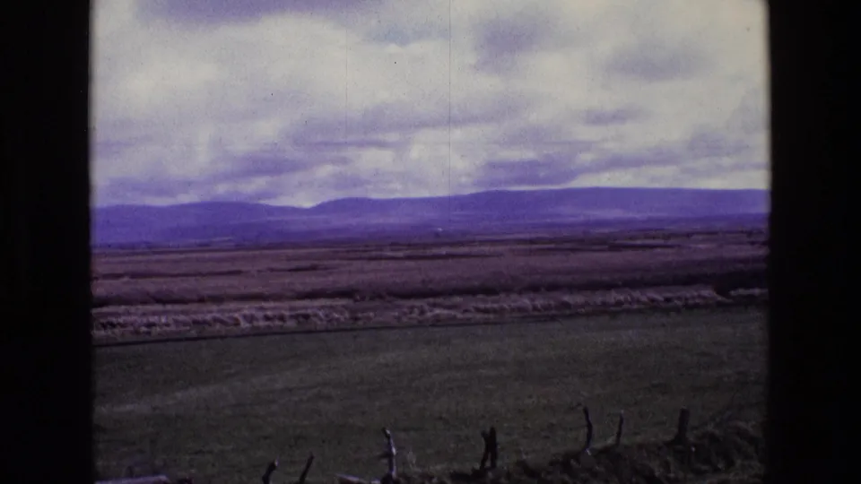 1969 Ireland frame with green fields, rough fence posts, distant hills, and a muted cloudy sky from a home-movie viewpoint.