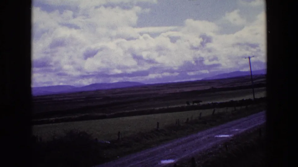 1969 Ireland home-movie frame showing a narrow rural road, roadside verge, fence line, open fields, and distant hills under large clouds.