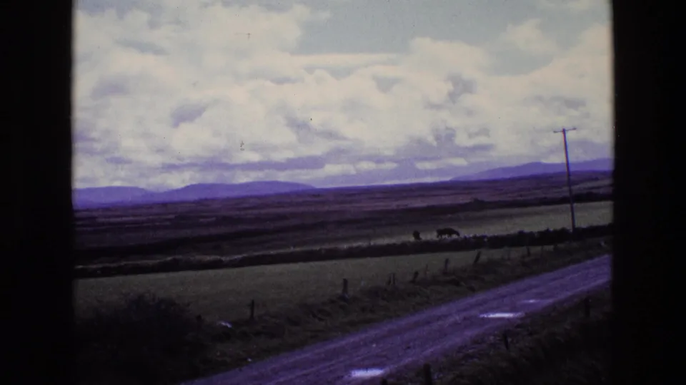 1969 Ireland 8mm view of a rural road, fields, distant hills, cloudy sky, and a utility pole framed by dark window edges.