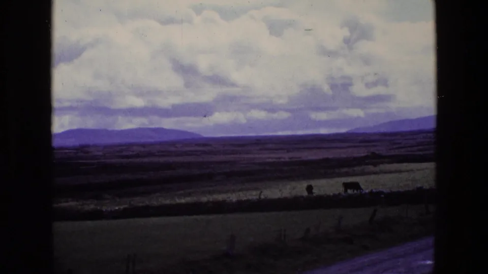 1969 Ireland countryside view with fields, low hills, a cloudy sky, and a dark vertical window frame crossing the shot.