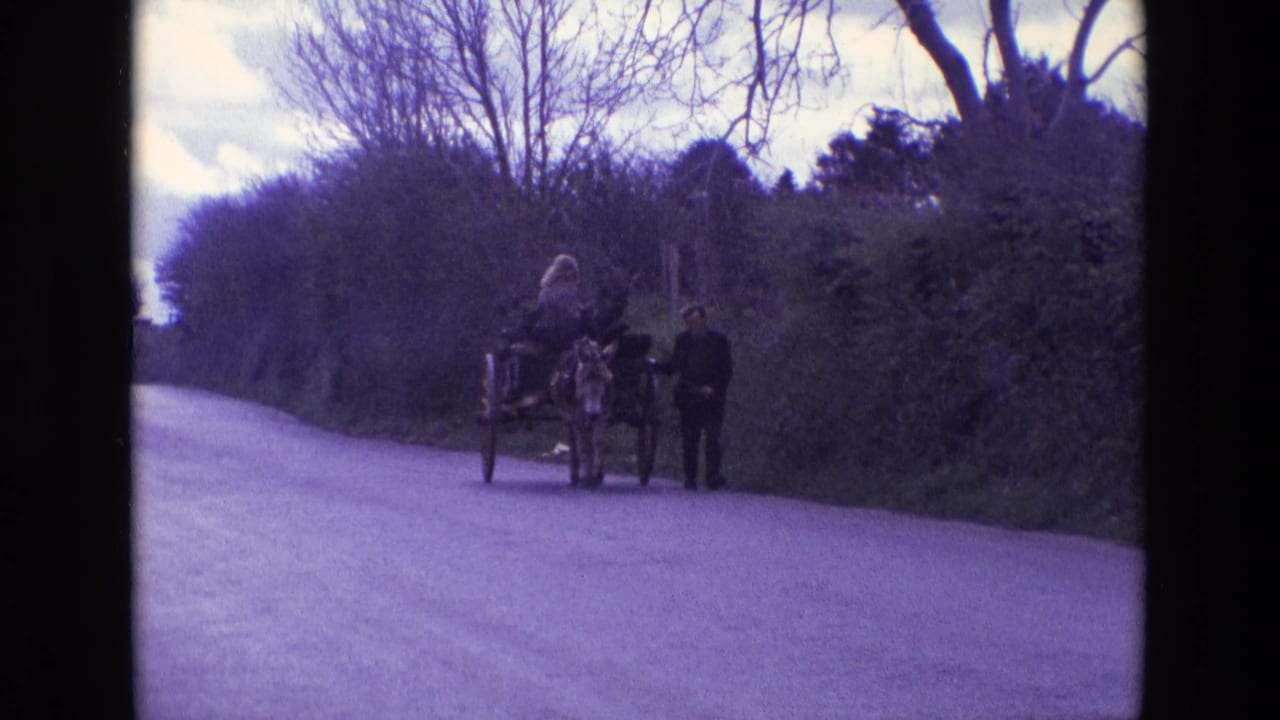 1969 Ireland home-movie frame showing a man leading a donkey cart with a woman seated on a quiet rural lane.