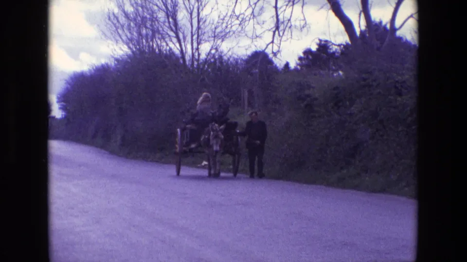 1969 Ireland home-movie frame showing a man leading a donkey cart with a woman seated on a quiet rural lane.
