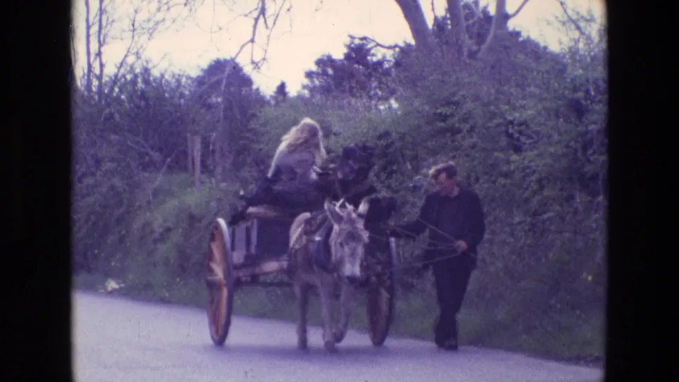 Wide 1969 Ireland home-movie frame showing a donkey pulling a wooden cart while a woman rides and a man walks alongside on a quiet rural road.
