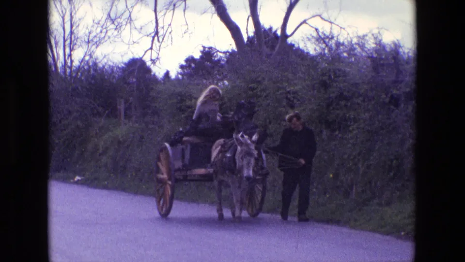 1969 Ireland 8mm frame showing the donkey cart farther along the road, with the seated woman, man at the reins, hedges, and trees in view.